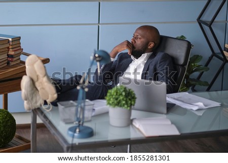 Relaxed worker finished work completed task taking break after hard workday putting head on hand. Thoughtful African American businessman looking out window sitting at office desk. Royalty-Free Stock Photo #1855281301