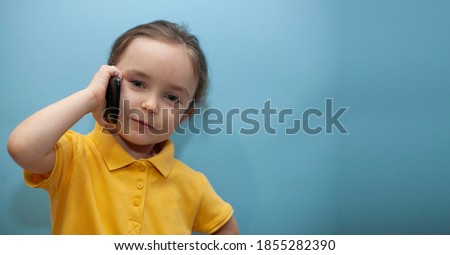 Little girl in a yellow T-shirt on a blue background. Child speaks on the phone and looks at the camera Royalty-Free Stock Photo #1855282390