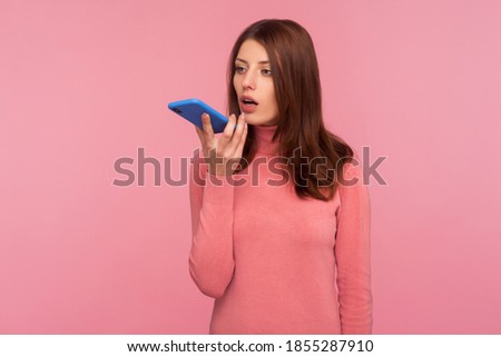 Business woman with brown hair in pink sweater holding mobile phone in hands and talking into load speaker, making voice note, voice assistant. Indoor studio shot isolated on pink background