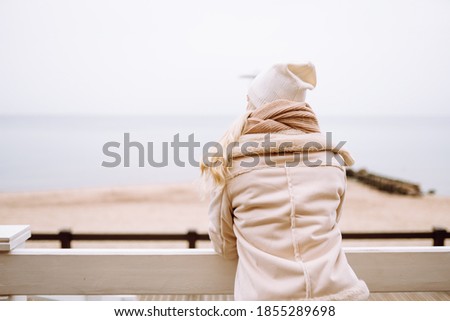 Back shot of blonde haired woman enjoy seaview at winter day. Female wearing a light jacket, hat, scarf and have hot beverage. Royalty-Free Stock Photo #1855289698