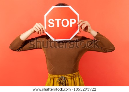 Scared anonymous woman hiding her face behind stop road sign, campaign against domestic violence, protection of women's rights. Indoor studio shot isolated on orange background