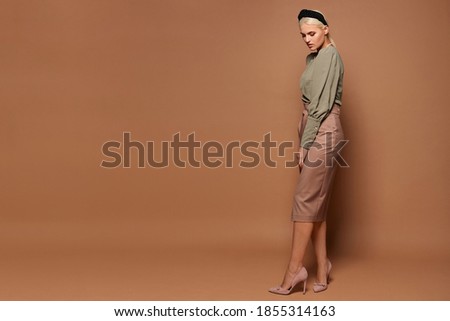Full-length studio shot of a model girl with blond hair wearing midi size skirt and blouse isolated at the beige background, side view