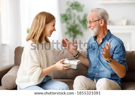 Excited young daughter smiling and giving gift box to cheerful aged man while sitting on couch during holiday celebration
