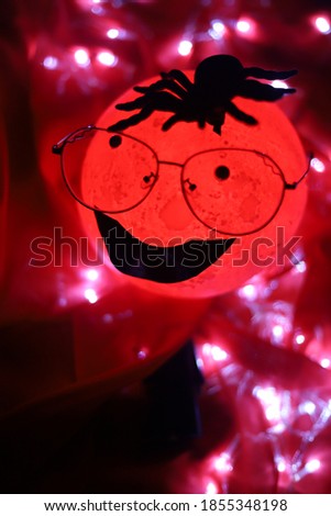 A very creepy cartoon moon shaped round light wearing glasses and a tarantula shaped spider over it's head smiling isolated on red background