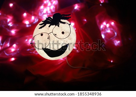 A very creepy cartoon moon shaped round white light wearing glasses looking strait at camera and a tarantula shaped spider over it's head smiling isolated on red background