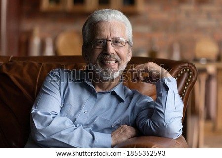 Head shot portrait smiling grey haired mature man wearing glasses sitting on cozy sofa at home, overjoyed happy senior male looking at camera, posing for photo, feeling positive, elderly generation