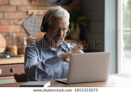 Close up mature man wearing headphones using laptop, making video call, sitting at table in kitchen, senior teacher mentor wearing glasses engaged online conference, recording webinar, teaching Royalty-Free Stock Photo #1855353073