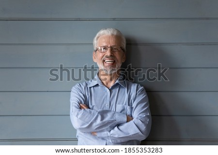 Head shot portrait laughing mature man wearing glasses standing with arms crossed on grey wooden wall background, overjoyed senior grandfather with healthy toothy smile looking at camera
