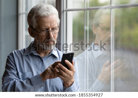 Close up serious mature man wearing glasses using phone, standing near window at home, lonely grandfather waiting for call from relatives or grandchildren, looking at smartphone screen Royalty-Free Stock Photo #1855354927