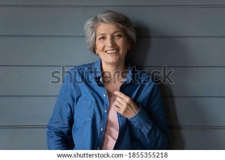 Head shot portrait smiling mature grey haired woman standing on grey wooden wall background, happy attractive senior older female looking at camera, posing for photo at home, retirement
