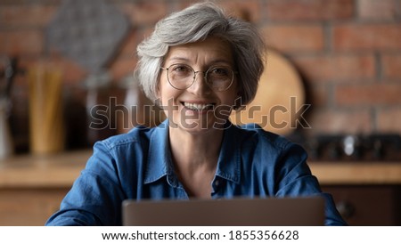Head shot portrait smiling mature woman wearing glasses using laptop, teacher mentor working online at home, happy confident senior businesswoman looking at camera, enjoying leisure time with device