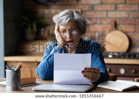 Unhappy mature wearing glasses reading bad news in letter, stressed grey haired female with open mouth looking at paper sheet, sitting at desk, shocked by negative message in correspondence Royalty-Free Stock Photo #1855356667