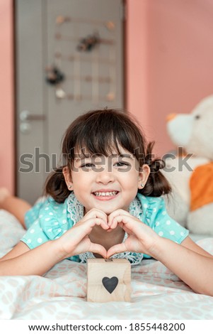 Little girl making sign heart gesture by fingers in bedroom.