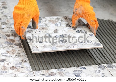 Worker's hands hold a tile and place it on floor smeared with glue. Process of laying ceramic tiles. Close up. Royalty-Free Stock Photo #1855470025