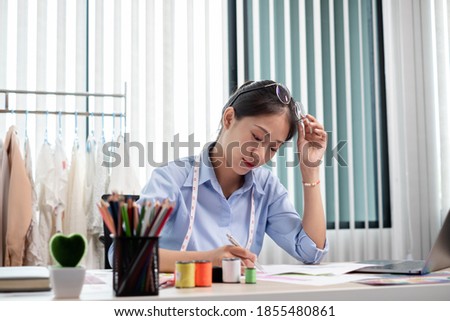Beautiful young Asian fashion tailor with textile sewing accessories and entrepreneur designer sketches are full of bright colors on the desk with a laptop for creative ideas in the studio.