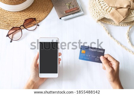 Hand holding credit card while using smart phone with travel accessories on bed. Holiday, technology and lifestyle, cashless society concept.