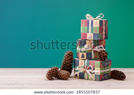 Gifts in vertical boxes, wrapped in paper with Christmas and New Year pictures, on light wooden table. Pine cones on green background.