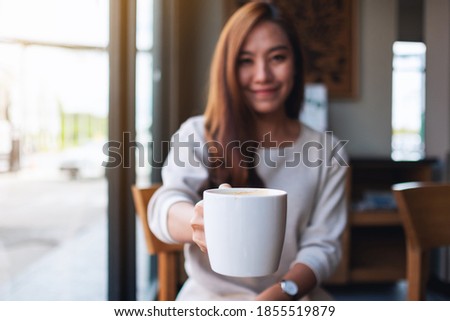 Closeup image of a beautiful young asian woman holding and giving a cup of hot coffee in cafe
