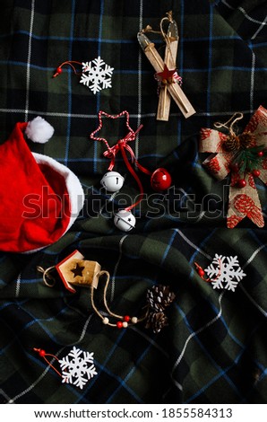 Scandinavian-style Christmas tree decorations are laid out on a green checkered blanket: bow, skis, house, snowflakes, etc. There is also a Santa Claus hat on the blanket.
