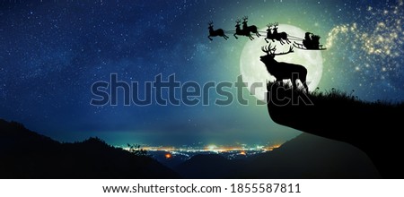 Silhouette of reindeer standing on the cliff to see Santa Claus flying on their reindeer over the full moon at night Christmas.