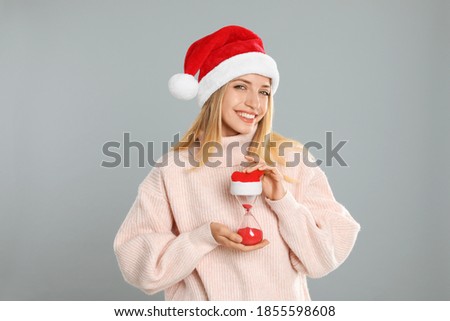 Woman in Santa hat with hourglass on grey background. Christmas countdown