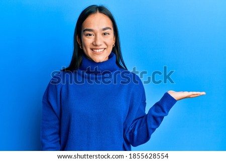 Young asian woman wearing casual winter sweater smiling cheerful presenting and pointing with palm of hand looking at the camera. 