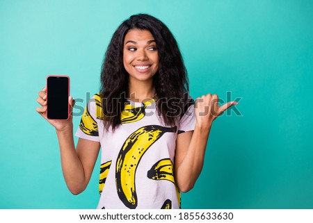 Photo of cute girl hold telephone screen empty space direct thumb look side wear banana print t-shirt isolated turquoise color background