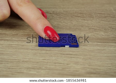 A blue memory card for a camera lies on a table and a woman's finger with a red manicure presses the card with her finger