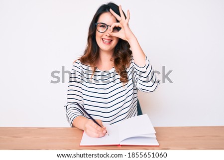 Young beautiful brunette woman writing book sitting on the table smiling happy doing ok sign with hand on eye looking through fingers 