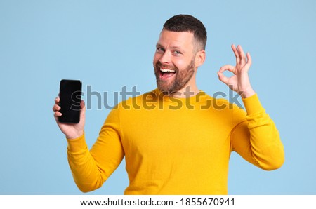 funny cheerful man in a yellow sweater with smartphone makes a gesture of approval on colored yellow background

