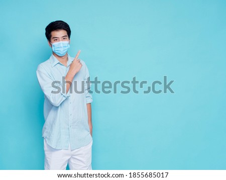 Portrait of handsome young man wearing medical mask in casual clothes pointing finger to space for text, on blue background. concept covid 19