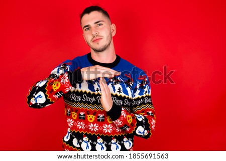 Young handsome Caucasian man wearing Christmas sweater against red wall, being upset showing a timeout gesture, needs stop, asks time for rest after hard work, demonstrates break hand sign