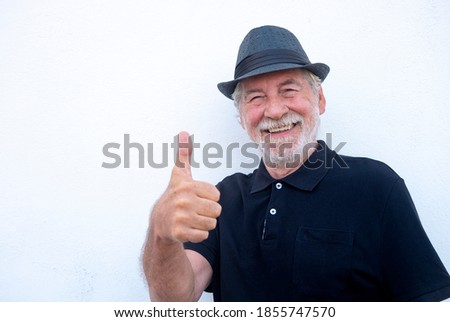 Portrait close-up of a smiling senior man with white beard on white background wearing a black hat and vest. Positive retiree person with thumb up