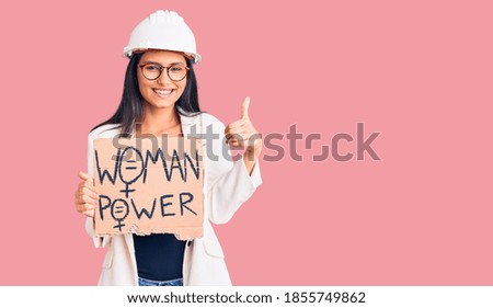 Young beautiful latin girl wearing architect hardhat holding woman power banner smiling happy and positive, thumb up doing excellent and approval sign 