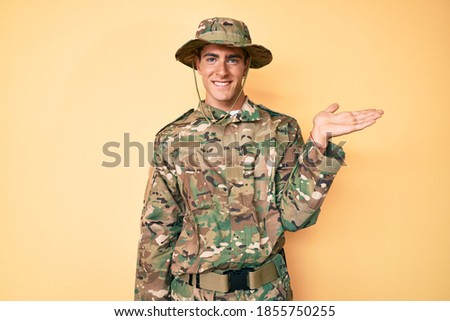 Young handsome man wearing camouflage army uniform smiling cheerful presenting and pointing with palm of hand looking at the camera. 