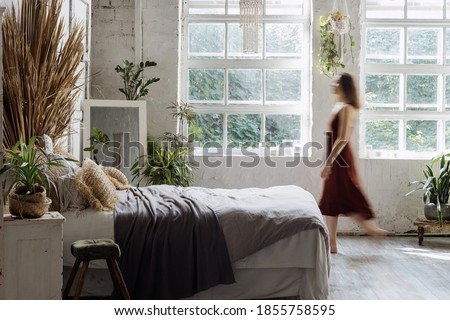 Blurred silhouette of young adult woman walking at comfortable bedroom with interior design in boho chic style. Lady in dress standing near cozy bed, enjoying morning at home