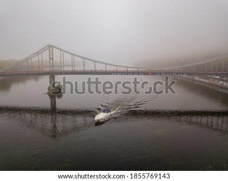 Aerial drone view. A motor boat sails under a footbridge on the Dnieper on a foggy morning.