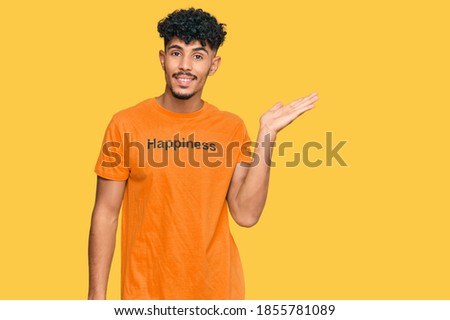Young arab man wearing tshirt with happiness word message smiling cheerful presenting and pointing with palm of hand looking at the camera. 