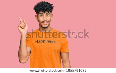 Young arab man wearing tshirt with happiness word message with a big smile on face, pointing with hand finger to the side looking at the camera. 
