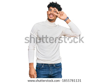 Young arab man wearing casual winter sweater doing peace symbol with fingers over face, smiling cheerful showing victory 