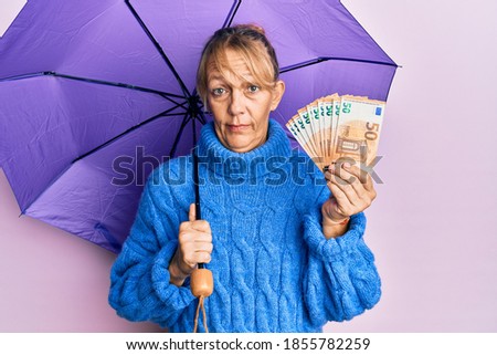 Middle age blonde woman holding umbrella and 50 euros banknotes clueless and confused expression. doubt concept. 