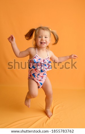 A little girl dressed in a swimsuit at the age of one and a half years is jumping or dancing. The girl is very happy. Picture taken in the studio on a yellow background.