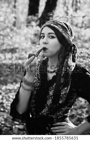 young gypsy woman smoking pipe in autumn forest, looking at camera, monochrome