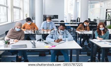 Modern Education And Studying During Pandemic Concept. Diverse group of international students sitting at desks in classroom at high school, wearing protective medical mask, writing in notebooks