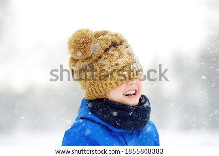 Funny little boy in blue winter clothes walks during a snowfall. Baby having fun while blizzard. Cute child wearing a warm hat low over his eyes. Outdoors winter activities for family with kids.