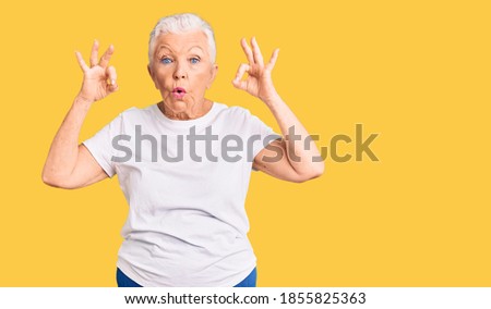 Senior beautiful woman with blue eyes and grey hair wearing casual white tshirt looking surprised and shocked doing ok approval symbol with fingers. crazy expression 