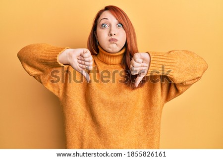 Beautiful redhead woman doing negative thumbs down gesture puffing cheeks with funny face. mouth inflated with air, catching air. 
