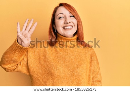 Beautiful redhead woman wearing casual winter sweater over yellow background showing and pointing up with fingers number four while smiling confident and happy. 