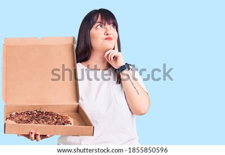Young plus size woman holding delivery pizza box serious face thinking about question with hand on chin, thoughtful about confusing idea 