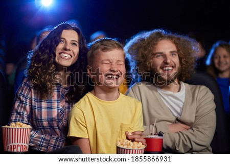 Selective focus of parents with curly hair enjoying time with son holding sparkling drink in cinema. Happy young family sitting in cinema, kid laughing with closed eyes, watching funny cartoon.